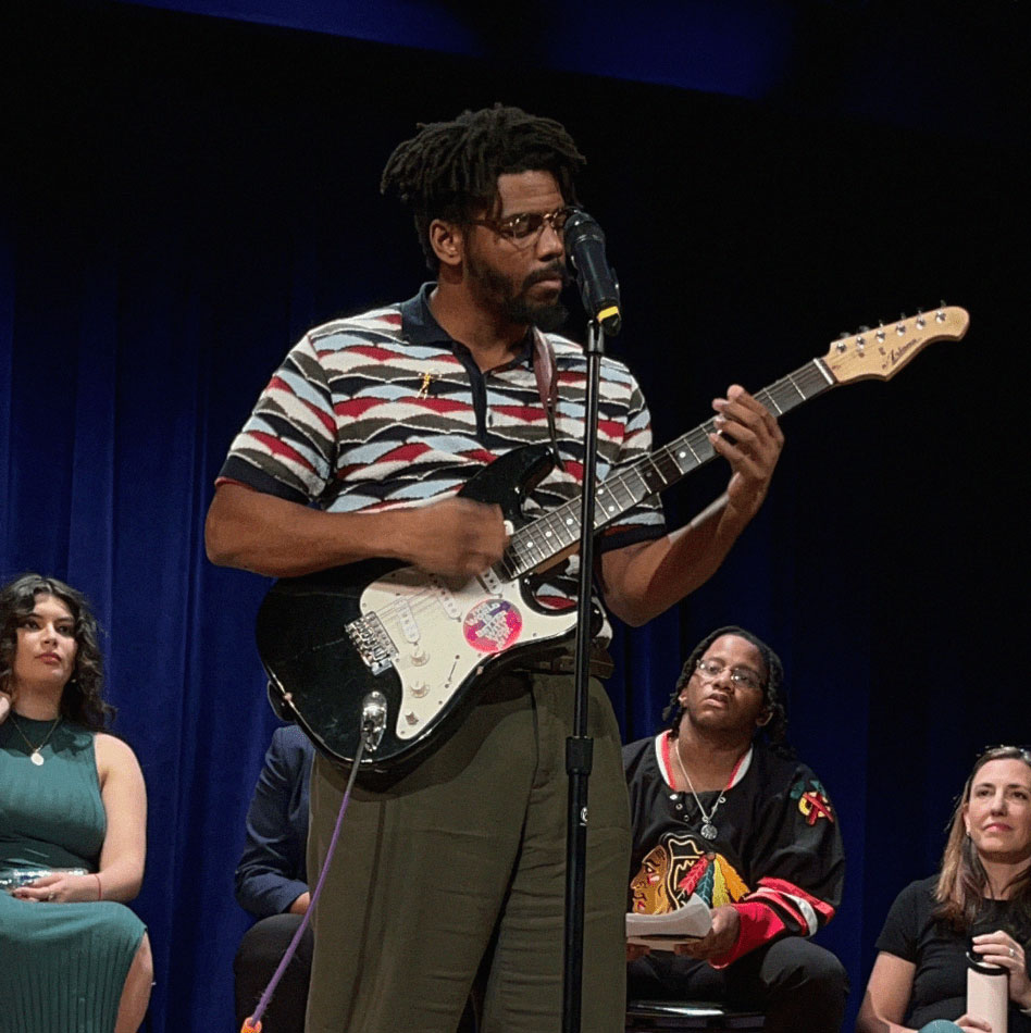 A man in a colorful shirt stands on stage in front of a microphone and plays a guitar. His guitar has a Society of Valued Minds sticker on it that says, "THE WORLD IS BETTER WITH YOU IN IT."