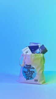 Background image of a Society of Valued Minds tote bag with a graphic reading "THE WORLD IS BETTER WITH YOU IN IT". The bag is overflowing with other merchandise, including a t-shirt, a pair of socks, a journal, and a "Mental Health Multi-Tool" resource. 